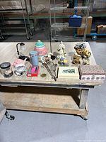 Photo showing miscellaneous home decor items and small appliances on a table in a warehouse setting, including figurines, cookie jar, lamp, fan, phone case, jewelry box, and photo frame.
