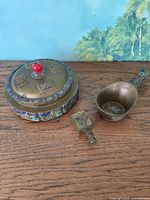 Three-piece oriental brass lot displayed on wooden surface against backdrop with palm trees, shows lidded container, scooping cup, and square box with handle.