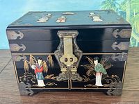 Front view of black lacquer jewelry box with brass lock and corner protectors, hand-painted Asian figures in red and green robes, and gold detailing of plants and landscape.