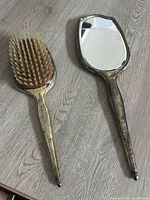 Photo showing the hand mirror and hairbrush side by side on a light wood floor. Mirror glass is cracked. Both have tarnished silver handles and backs.