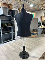 Photo showing front view of the black fabric shirt mannequin on black circular base with finial top in a warehouse setting.