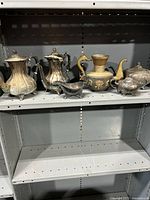 Shelf view of five tea and coffee pots including silver and brass-tone pieces with ornate handles and lids.