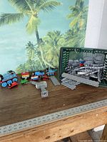 Overall image showing multiple Thomas the Train toy trains and a crate filled with numerous grey track pieces on a wooden table with a tropical hemmed wall background.