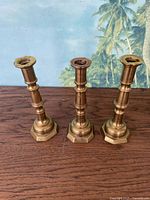 Front view of three brass candlesticks with hexagonal bases and decorative shafts on wooden surface against palm tree background.