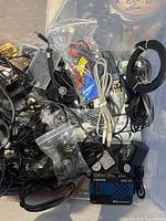 Top view of large tote showing a mixture of electronic cables including HDMI, USB, audio, and power adapters along with some car parts and remotes in plastic bags.