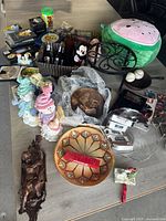 Wide view of lot on table showing watermelon plush stool, Mickey Mouse figure, brown horse candy dish, decorative pottery plate and various figurines, toys, glass items and collectibles