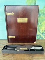 Wooden bible box with visible brass hinges and engraved brass plate on front cover, showing wear and corner damage. Accompanied by a black-handled cake cutting set in fitted box.
