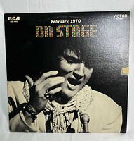 Front cover of Elvis 'On Stage - February, 1970' vinyl record showing Elvis wearing a white stage costume with ruffled scarf, slight tape damage and minor cover wear visible.