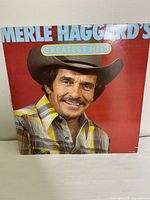 Front cover of Merle Haggard's Greatest Hits vinyl album, showing his portrait in a hat and plaid shirt with red background and album title.