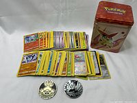 Stack of Pokemon cards, 2 plastic collectible coins (gold and silver tone), and Pokemon collector tin with Victini design