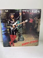 Front cover of the 'Street Songs' vinyl album showing Rick James standing on a street at night next to a lamp post wearing black leather clothes and red boots.
