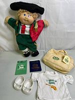 Full view of the doll standing with the beige tote bag, white doll shoes, white printed shirt, and two booklets laid out in front on a flat surface