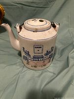 Front view of large white porcelain teapot with blue painted buildings and floral designs, rope handles visible