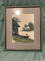 Framed signed fabric artwork featuring a river scene with stilt houses and a person paddling a boat.