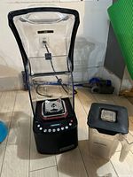 Black Blendtec blender base with clear plastic sound enclosure on wooden floor, showing front with control buttons.