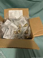 Open cardboard box filled with multiple small plastic bags each containing brass K1 connection fittings for pipes.