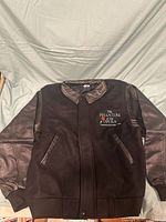 Front view of dark brown jacket with faux leather collar and sleeve accents. Embroidered logo on left chest with red rose and The Phantom of the Opera text.