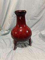 Front angle view of a red glazed Chinese style vase, showing the bulbous body and neck with three supporting legs.