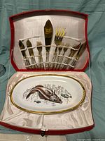 Fish cutlery set including knives and forks with ornate handles in a red box, and accompanying oval ceramic platter showing a painted fish.
