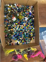 Overhead view of cardboard box filled mostly with small-to-medium mixed color glass marbles, plus a few large marbles