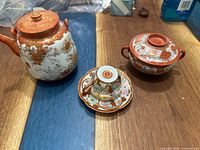 Three-piece Japanese Kutani porcelain tea set including teapot, covered bowl, and teacup with saucer, all decorated in orange, gold, and white with traditional patterns.