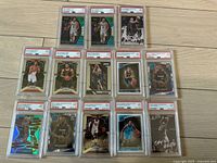 Overview of all 13 basketball cards graded by PSA in protective slabs laid out in three rows on a wooden floor.