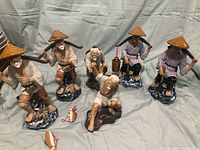 Wide shot of six Chinese Shiwan fisherman/farmer porcelain statues arranged in group showing different poses and styles with two ceramic fish figurines on the fabric background.