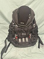 Front view of black Oakley Kitchen Sink backpack showing zippered compartments, adjustable side straps, and Oakley logo on front top.
