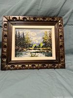 Framed oil painting depicting a landscape with trees, a reflective body of water, and mountains in the background.