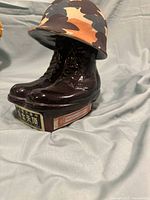 Close-up side view of ceramic military boots decanter with helmet lid showing the base with Chinese characters and label, on grey cloth background.