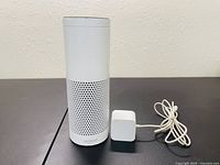 Front view of white Amazon Alexa Echo smart speaker standing upright beside its white power adapter with attached cord on neutral surface and wall background.