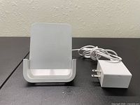 Front view of Apple MagSafe wireless charger stand with Apple power adapter. Shows vertical charger surface and coiled white cable attached to power adapter.