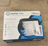 Front of Ooma Telo boxed device, showing key features and branding with shipping labels partly covering the box front.