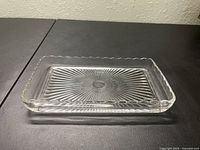 Clear rectangular glass tray dish viewed from the side showing thickness and scalloped edge detail on dark surface.