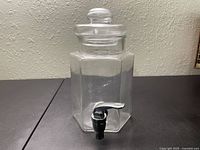 Clear glass lemonade dispenser with hexagonal shape, glass lid, and black spigot, shown from front angle