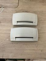 Two Zebra Full Cutter 102215G-003 components shown on a wooden surface; both white plastic parts with label cutter slots and vent markings visible; light wear noted.