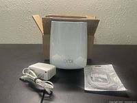 Front view of the white Netgear Orbi Router with power adapter and sealed Ethernet cable in front and an open brown cardboard box behind.