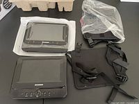 Photo showing main DVD player unit with screen and front control buttons, smaller secondary screen wrapped in bubble wrap, and two black mounting straps