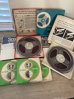Photo showing multiple Scotch brand open reel magnetic tapes, including reels on plastic spools, original boxes, Irish brand paper sleeves, and various product literature sheets with product instructions and claims.