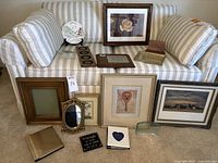 Overview image showing large brown empty frame, several framed artworks, decorative plate on stand, old age plaque, photo albums, and small mirror on a striped couch
