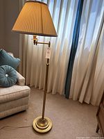 Full view of the brass floor lamp by a beige upholstered armchair and window with curtains.