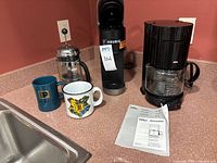 Bodum French press, two coffee mugs, Keurig coffee maker, Braun 10 cup coffee maker with manual on kitchen counter near sink.