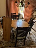 Full view of round wooden pedestal table with two leaves leaning against it, surrounded by six chairs with light-colored upholstered seats.