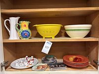 Shelf with assorted kitchenware: white and floral pitchers, bright yellow plastic colander with green stripes, beige mixing bowl with green stripe, various decorative items and ceramics