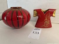 Both the red lusterware pot and the small porcelain robe with gold Arabic calligraphy shown together on a flat surface in front of a white background.