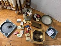 Full overview of all items including ashtrays, metal cricket figurine, cigarette case, and matchbooks in a tin.
