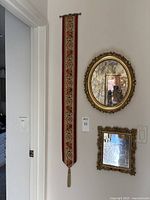 The entire lot showing an oval gold framed mirror, a smaller rectangular gold framed mirror below it, and an embroidered bell pull hanging vertically beside them.