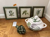 Three framed herb prints and Alan Umbro soup tureen with porcelain artichoke figurine on wooden floor.