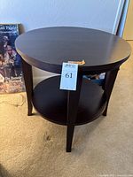 Dark wood round side table with two levels shown from front view with tag attached to front leg and artwork in background.