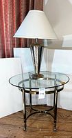 Full view of the hammered metal lamp with white lampshade sitting on the round glass top brass frame side table.